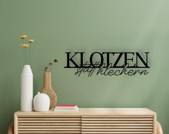 May include: Black metal wall art with the German phrase "KLOTZEN statt kleckern" displayed on a green wall. The art is above a wooden cabinet with vases and flowers.