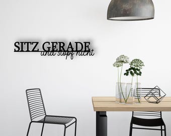 May include: A modern interior scene featuring a black metal chair, a wooden table, and a decorative metal sign on the wall that reads "SITZ GERADE und stopf nicht". The scene is illuminated by a hanging lamp and includes floral arrangements.