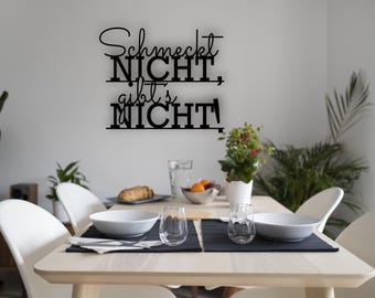 May include: A dining room scene featuring a wooden table set for a meal. Above the table, a black metal wall art piece displays the German phrase "Schmeckt NICHT, gibt's NICHT." The table is set with white bowls, glasses, and a small plant.
