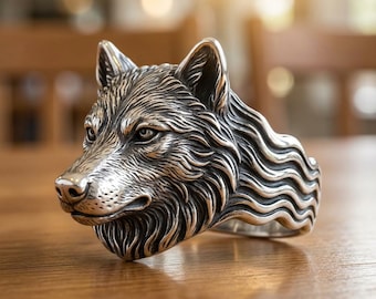 May include: A silver-colored wolf head ring. The ring features detailed fur texture and a realistic wolf face. The ring is set on a wooden surface, with a blurred background.