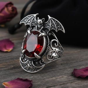 Könnte beinhalten: Silberner Ring mit einem großen, ovalen, roten Edelstein. Der Ring ist mit einer Fledermaus und Monddetails verziert. Der Ring hat aufwendige Filigranmuster. Der Ring ist im Gothic-Stil.