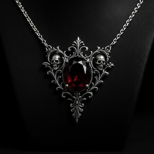 Puede incluir: Collar con colgante plateado con una gema roja oscura ovalada y facetada. El colgante tiene un diseño gótico adornado con dos pequeñas calaveras y un intrincado trabajo de volutas. El collar se muestra sobre un fondo negro.