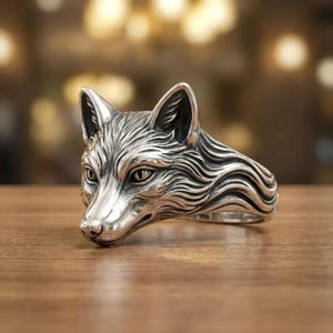 May include: A silver-colored ring featuring a detailed fox head design. The ring showcases the fox's face, ears, and fur texture. The ring is made of a shiny, metallic material and is set against a blurred background.
