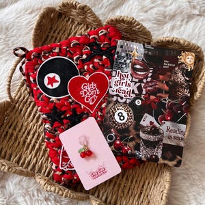 May include: A red and black crocheted bag with patches, including a record, a heart with "Girls Don't Cry," and stars. A collage with the text "hot girls read" and "Reading is sexy" is next to the bag. A pink card with cherry earrings is also present.