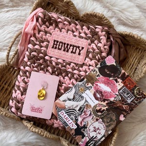 May include: A pink and brown crocheted bag with a rectangular patch reading HOWDY, a small gold cowboy hat pin on a pink card, and a rectangular sticker sheet featuring various western-themed graphics, lip prints, and the text DARLIN'.
