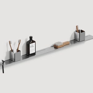 Puede incluir: Estante de baño minimalista de acero inoxidable con varios artículos de aseo. Cuenta con dos porta cepillos de dientes, una botella, un cepillo de madera, un peine y una maquinilla de afeitar. El estante está montado en una pared blanca.