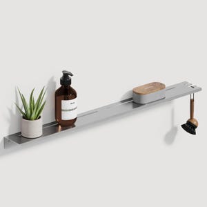 Puede incluir: Una estantería de pared larga de metal plateado cepillado con una pequeña suculenta verde en una maceta gris, un dispensador de jabón de vidrio ámbar marrón con etiqueta blanca, un recipiente gris rectangular con tapa de madera y un cepillo.