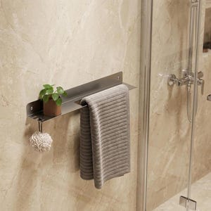 May include: A matte gray metal wall-mounted shelf featuring a flat top surface, a horizontal bar for hanging towels, and a small hook on the left side. A small green potted plant sits on the shelf, and a gray textured towel hangs from the bar.