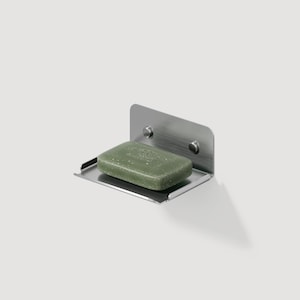 May include: A stainless steel soap dish with a rectangular green bar of soap. The dish is mounted on a white wall and has a small shelf to hold the soap. The soap dish is attached to the wall with two screws.
