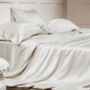 May include: A set of off-white silk bedding featuring a flat sheet, a fitted sheet, and two matching pillowcases. The fabric has a smooth, lustrous finish that drapes over the bed, creating soft folds and highlights across the surface of the material.