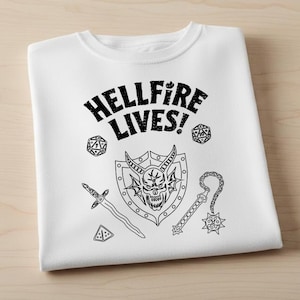 Pode incluir: Camiseta branca com um design gráfico preto. O design inclui o texto "HELLFIRE LIVES!" acima de um escudo com uma caveira, espada, maça e dados. A camiseta está dobrada em uma superfície de madeira.