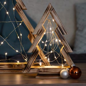 Op de afbeelding: Twee houten kerstboomversieringen met warmwitte lichtslingers aan de binnenkant. De bomen staan op een houten oppervlak met kerstversieringen ervoor. Op de achtergrond een gouden kaars.