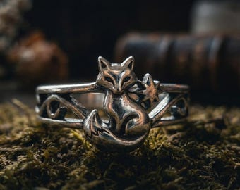 May include: A silver ring featuring a fox and star design. The ring has an intricate openwork band with a fox figure at the center, accompanied by a star. The ring is set against a mossy background.