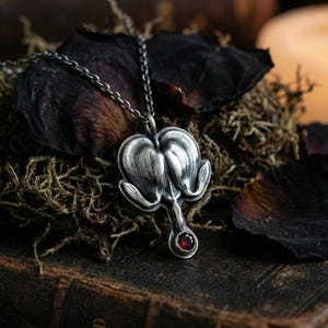 May include: A silver-toned pendant necklace featuring a stylized heart-shaped design with a small, round, red gemstone at the bottom. The pendant hangs from a delicate chain. The necklace rests on a bed of moss and dried rose petals.