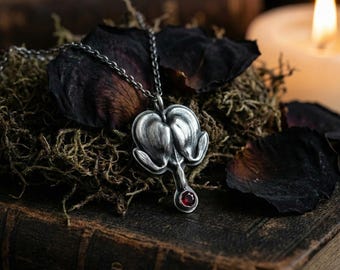 May include: A silver-toned pendant necklace featuring a stylized heart-shaped design with a small, round, red gemstone at the bottom. The pendant hangs from a delicate chain. The necklace rests on a bed of moss and dried rose petals.