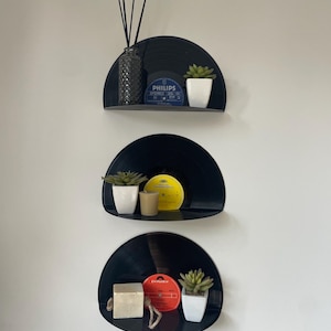 Peut inclure: Trois étagères murales en forme de demi-cercle fabriquées à partir de disques vinyles noirs. Chaque étagère supporte de petits objets décoratifs, dont des pots blancs avec des plantes succulentes, un diffuseur et une bougie.