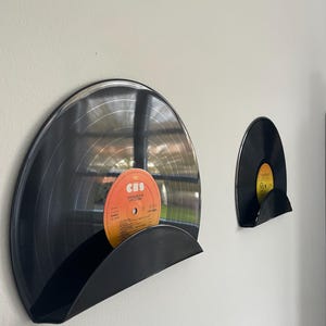 May include: Two black vinyl records mounted on a wall using curved black holders. The larger record features a circular orange label with the text CBS and Le Disque D'or Joe Dassin, while the smaller record has a yellow label.
