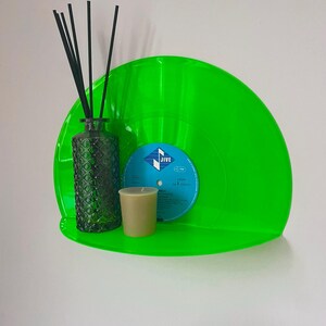 Peut inclure: Une étagère murale semi-circulaire vert néon vif fabriquée à partir d'un disque vinyle recyclé. L'étagère supporte un flacon diffuseur en verre texturé vert foncé avec des bâtonnets noirs et une petite bougie beige. L'étiquette est bleue avec JIVE.