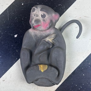 May include: A gray monkey figurine with pink cheeks and a red tongue, holding a banana. The monkey has a gold-colored patch on its lap and a curved tail. The figurine is sitting on a black and white striped surface.