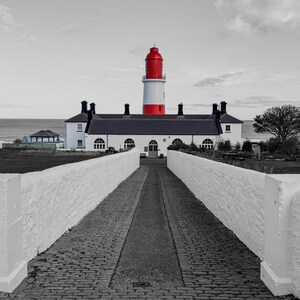 May include: A black and white image of a lighthouse with a red top and white body. A brick pathway leads to the lighthouse, bordered by white walls. The sky is grey and overcast.