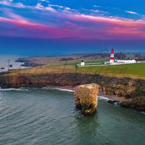 May include: Coastal landscape with a red and white lighthouse on a cliff, under a colourful sunset. The image shows the sea, rock formations, and green fields, typical of a British coastal scene.