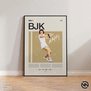 May include: A framed poster featuring a tennis player in action, wearing a white outfit with blue accents. The poster includes the initials "BJK", a signature, and text at the bottom. The background is tan with vertical white stripes.