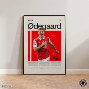 May include: A framed poster featuring a soccer player in a red and white jersey. The poster has a red background with the player's image and the name "Ødegaard" in large black letters. Includes the text "es BETTER" and a signature.