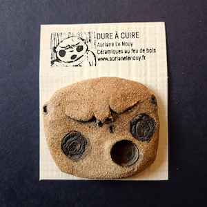 Peut inclure: Une pièce en céramique marron faite à la main, avec un design en forme de visage, comprenant deux impressions sombres et circulaires en forme d'yeux et une ouverture plus grande, circulaire en forme de bouche. Une carte avec du texte et une illustration de dessin animé l'accompagne.