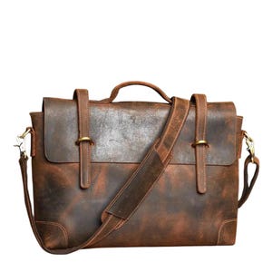 May include: A brown leather messenger bag with a flap closure, shoulder strap, and a top handle. The bag features brass-colored hardware and a distressed finish, giving it a vintage look. The bag is approximately 14 inches wide and 11 inches tall.