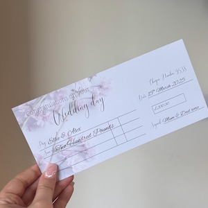May include: A white wedding cheque with floral accents and the text "Congratulations on your Wedding day." The cheque is made out to Ellie & Alex for Two Hundred Pounds, dated 15th March 2025, with cheque number 9538.