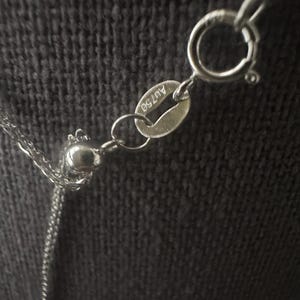 May include: Close-up of a silver necklace with a delicate chain and a round clasp. The necklace has a small tag with the marking "AU750". The background is a dark, textured fabric.