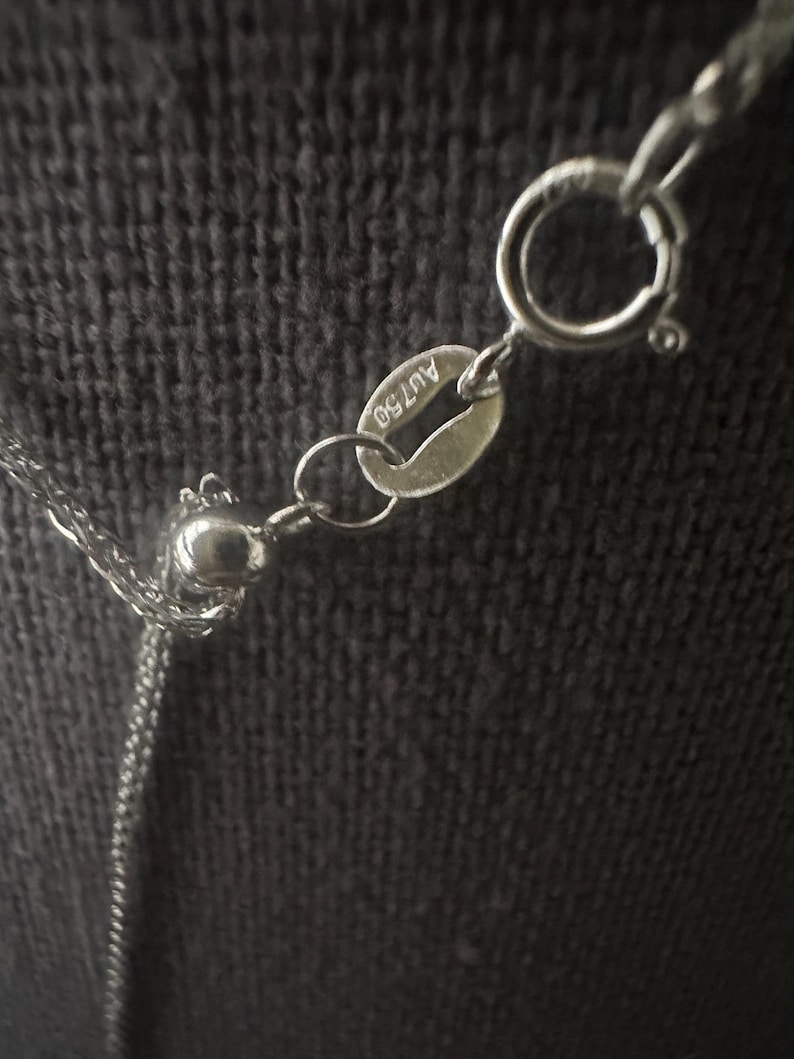 May include: Close-up of a silver necklace with a delicate chain and a round clasp. The necklace has a small tag with the marking "AU750". The background is a dark, textured fabric.