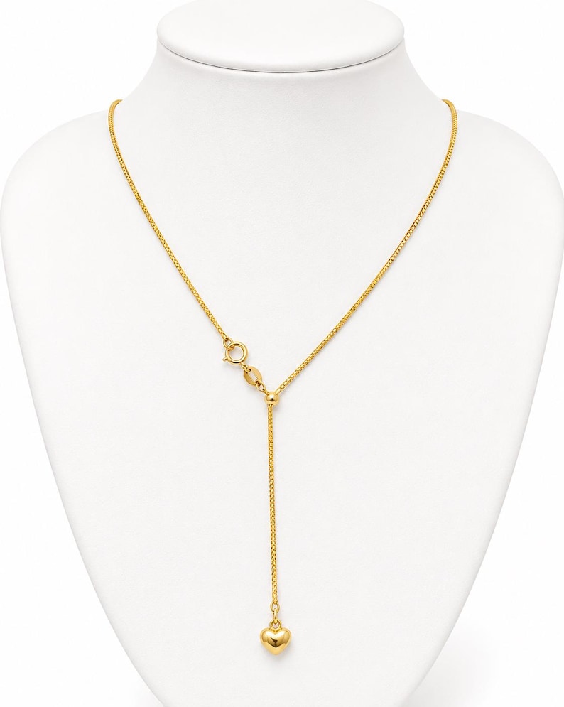 May include: A gold-tone necklace featuring a delicate chain with a sliding bead mechanism that allows for adjustable length. The chain terminates in a small, smooth, polished gold-tone heart charm suspended from the center of the Y-shaped design.