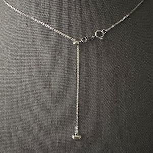 May include: A silver-toned necklace with a delicate chain and a small heart pendant. The necklace features an adjustable design with a sliding bead and a lobster clasp closure. The background is a neutral gray fabric.