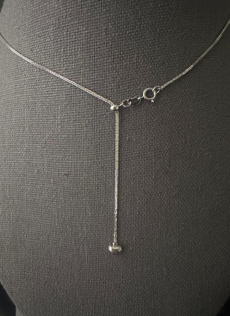 May include: A silver-toned necklace with a delicate chain and a small heart pendant. The necklace features an adjustable design with a sliding bead and a lobster clasp closure. The background is a neutral gray fabric.