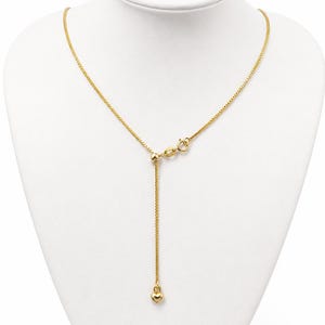 May include: A gold-toned box chain necklace featuring an adjustable sliding bead mechanism and a small, smooth heart-shaped charm dangling from the end of the chain. The necklace includes a circular spring ring clasp for secure fastening.