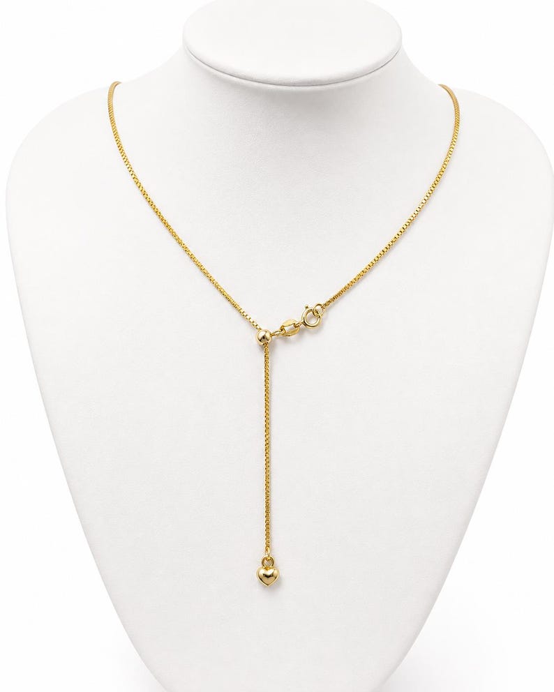 May include: A gold-toned box chain necklace featuring an adjustable sliding bead mechanism and a small, smooth heart-shaped charm dangling from the end of the chain. The necklace includes a circular spring ring clasp for secure fastening.