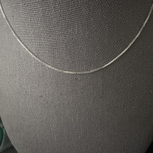 May include: A delicate silver chain necklace displayed against a gray backdrop. The chain is thin and appears to be made of small, interconnected links. The necklace is simple and elegant, suitable for everyday wear or layering with other jewelry.