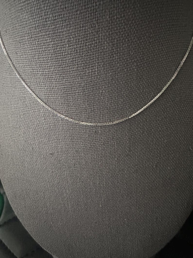 May include: A delicate silver chain necklace displayed against a gray backdrop. The chain is thin and appears to be made of small, interconnected links. The necklace is simple and elegant, suitable for everyday wear or layering with other jewelry.