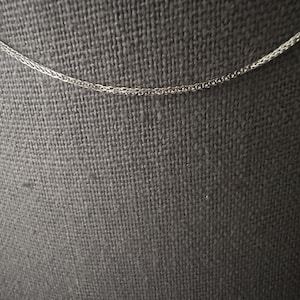 May include: A delicate silver-colored chain necklace. The chain has a wheat or herringbone pattern, with a fine, woven texture. The background is a dark gray fabric, providing contrast to the bright chain.