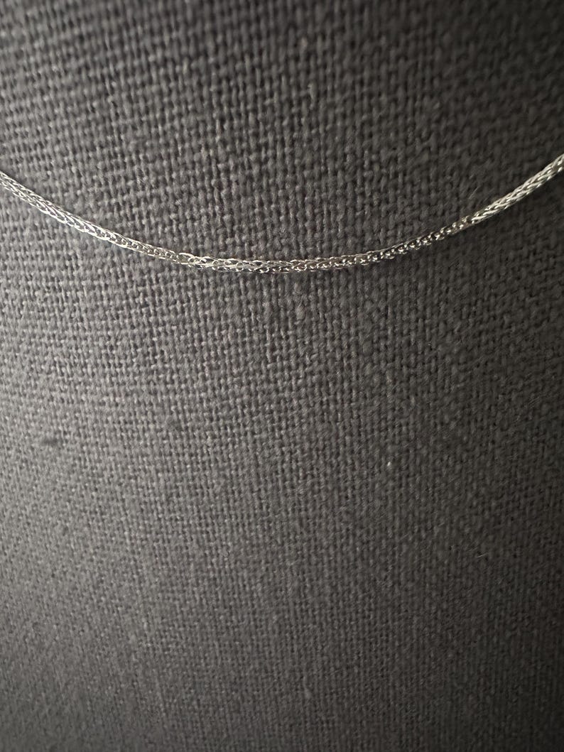 May include: A delicate silver-colored chain necklace. The chain has a wheat or herringbone pattern, with a fine, woven texture. The background is a dark gray fabric, providing contrast to the bright chain.