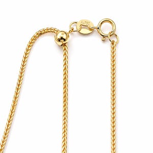 May include: A gold wheat chain necklace featuring a sliding adjustable bead mechanism and a spring ring clasp. A small oval tag attached to the clasp is clearly engraved with the text Au750, indicating the gold purity of the metal chain.