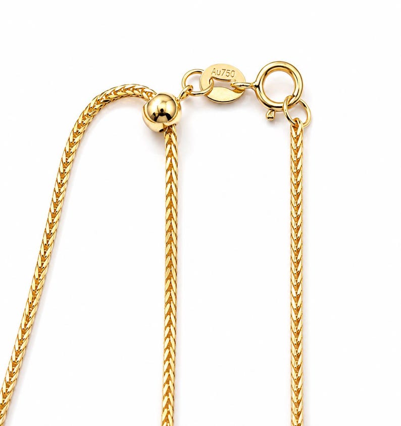 May include: A gold wheat chain necklace featuring a sliding adjustable bead mechanism and a spring ring clasp. A small oval tag attached to the clasp is clearly engraved with the text Au750, indicating the gold purity of the metal chain.