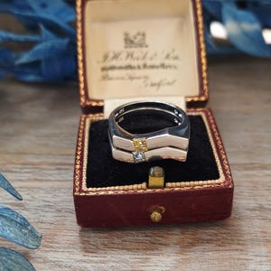 May include: A silver ring with a distinctive two-band design, featuring a small yellow and clear square stone. The ring is presented in a vintage red jewellery box with a black velvet interior, on a wooden surface.