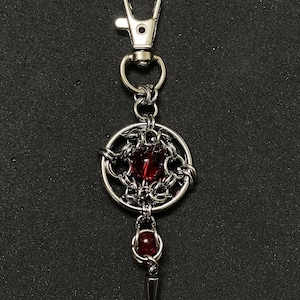 Może przedstawiać: Srebrny brelok do kluczy z ozdobną zawieszką. Zawieszka posiada centralny, okrągły, ciemnoczerwony kamień szlachetny, otoczony misterną metaloplastyką. Mniejszy czerwony kamień wisi poniżej, ze srebrnym kolcem na dole.
