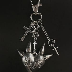 May include: A silver-toned metal keyring featuring a heart-shaped charm adorned with three prominent spikes and circular accents. The charm hangs from a lobster clasp via a series of small chains, decorated with two additional small cross pendants.