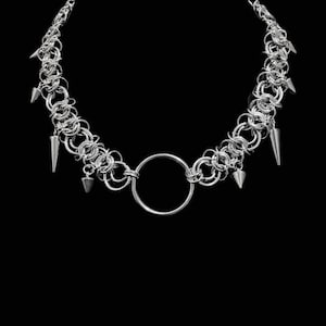 Op de afbeelding: Een zilverkleurige maliënkolderketting gemaakt van in elkaar grijpende metalen ringen. Het ontwerp heeft een opvallende cirkelvormige ring in het midden en verschillende puntige, kegelvormige stekels die aan de schakels hangen.