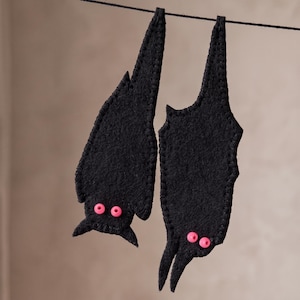 Peut inclure: Deux ornements chauve-souris en feutrine noire suspendus à un fil noir fin. Chaque chauve-souris présente un bord cousu et deux petites perles circulaires rose vif en guise d'yeux. Elles sont orientées verticalement, l'une plus basse.