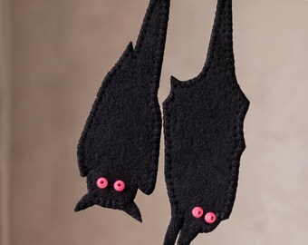 Könnte beinhalten: Zwei Fledermaus-Ornamente aus schwarzem Filz, die an einer dünnen schwarzen Schnur hängen. Jede Fledermaus hat einen genähten Rand und zwei kleine, leuchtend rosa runde Perlen als Augen. Die Fledermäuse hängen vertikal, eine etwas tiefer.