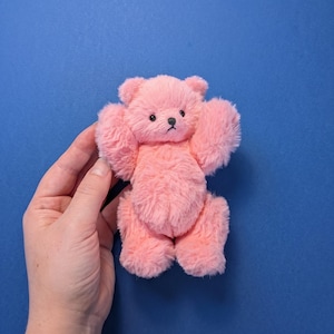 Peut inclure: Un petit ours en peluche avec une fourrure douce rose clair. L'ours présente des oreilles arrondies, un petit nez noir et des yeux ronds foncés. Ses bras sont levés vers le haut et ses jambes sont positionnées vers le bas en position assise.